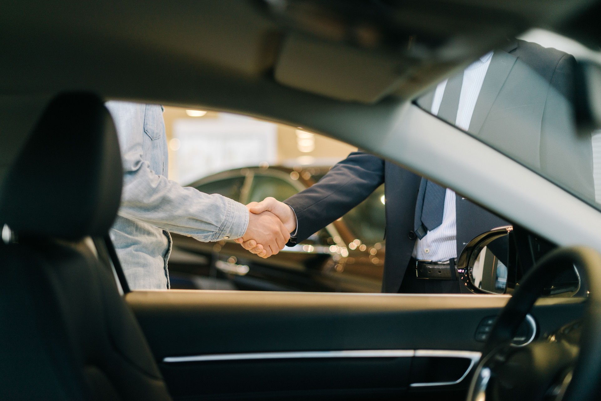 Buyer of car shaking hands with seller in auto dealership, view from interior of car.