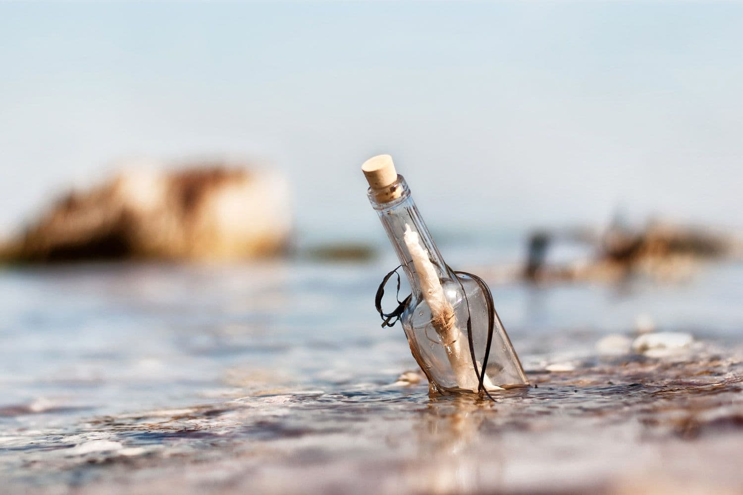 Corked glass bottle containing a rolled message rests in shallow water on a sandy beach.